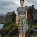A woman stands outdoors in front of a stone castle and moorland landscape, wearing a fitted gray short-sleeve crop top and a patterned wrap-style skirt with blue, green, beige, and gray tones.