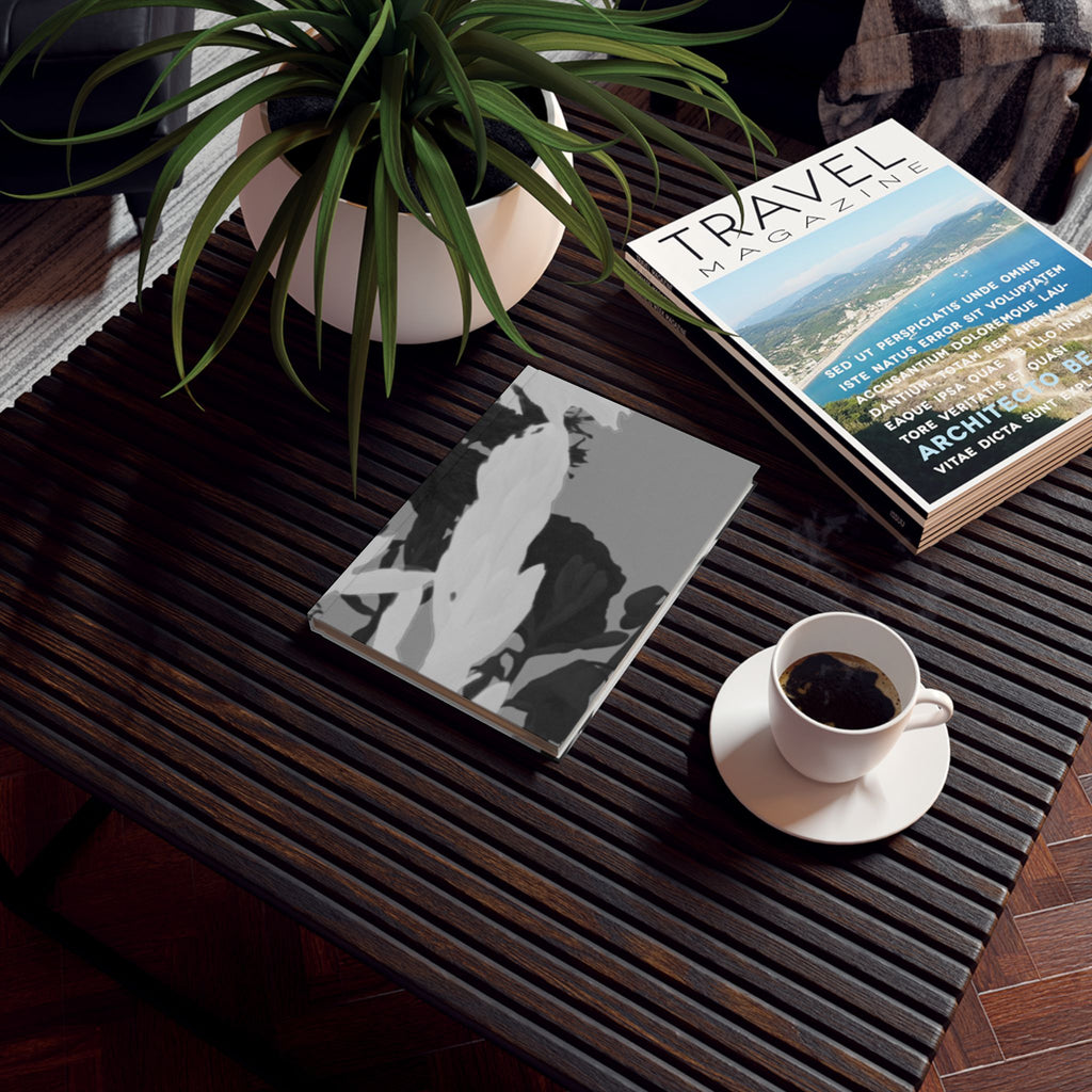 A styled tabletop scene with a dark slatted coffee table holding a potted green plant, a stack of magazines including one titled Travel Magazine, a gray-and-white patterned notebook or book, and a white cup of black coffee on a saucer.