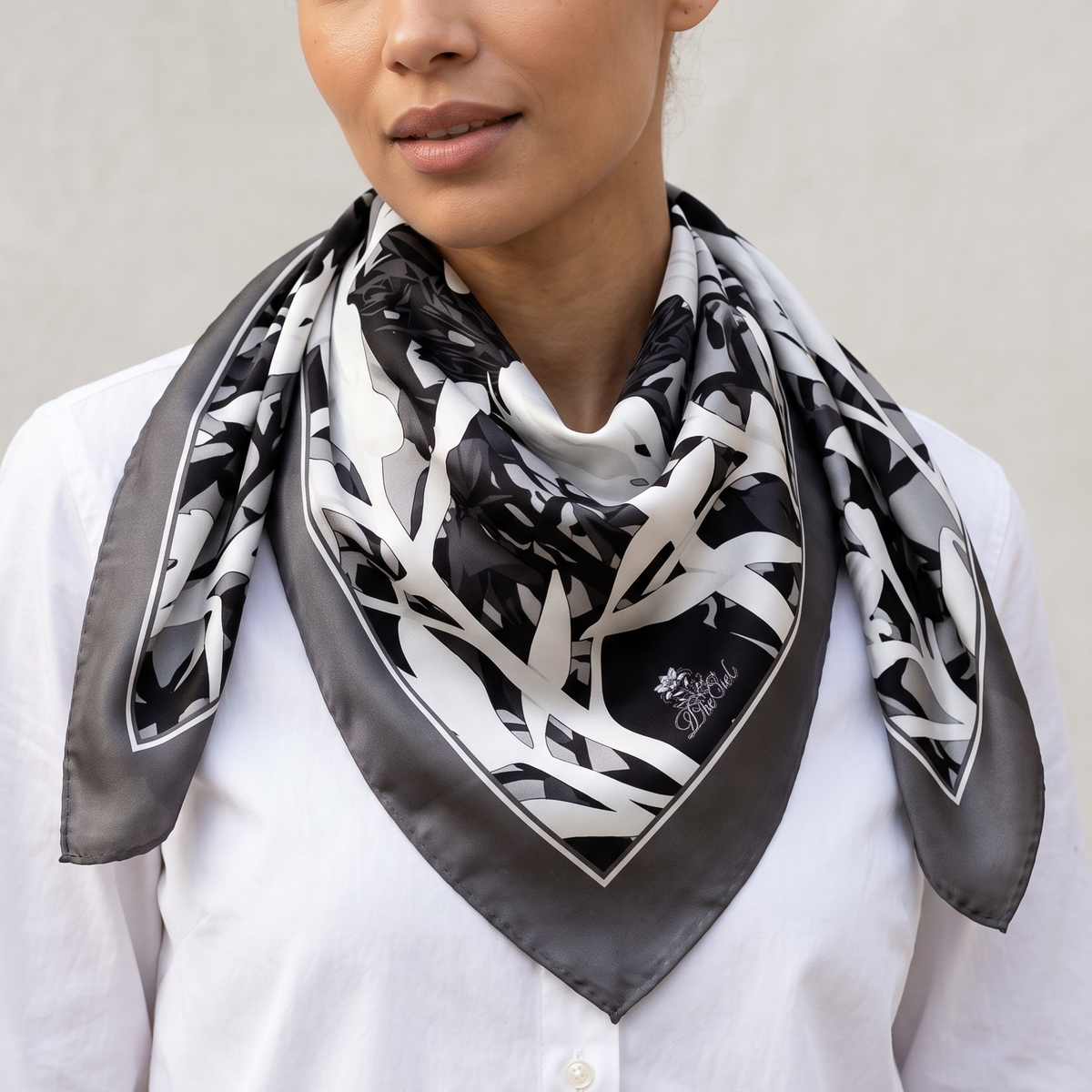 A woman wearing a black, white, and gray patterned silk scarf with a satin border, styled around her neck over a white button-down shirt against a neutral gray wall.