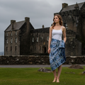 A woman stands barefoot on a grassy lawn in front of a large stone castle, wearing a white camisole top and a blue patterned sarong skirt.
