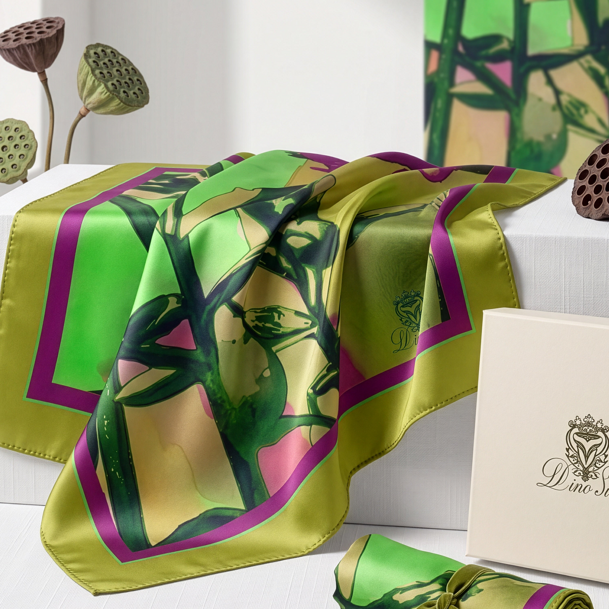 A silk scarf with a green and pink botanical print draped over a white display block, alongside its cream-colored presentation box and a rolled section with a ribbon. A small card displays product details, and decorative lotus pods and a purple flower sit in the background.