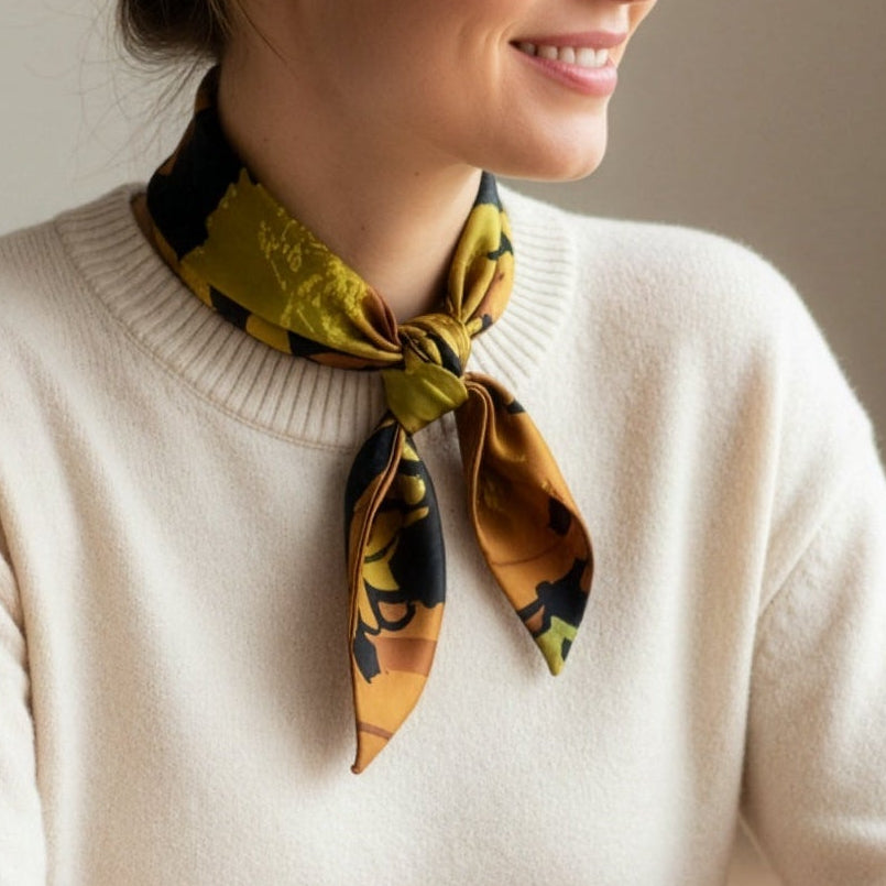 A woman with her hair in a bun wearing a cream knit sweater and a patterned silk neck scarf, looking to the side in a softly lit indoor setting.