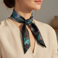 A woman wearing a cream-colored wrap blouse and a dark patterned silk neck scarf with teal and black floral motifs, photographed in warm natural light.