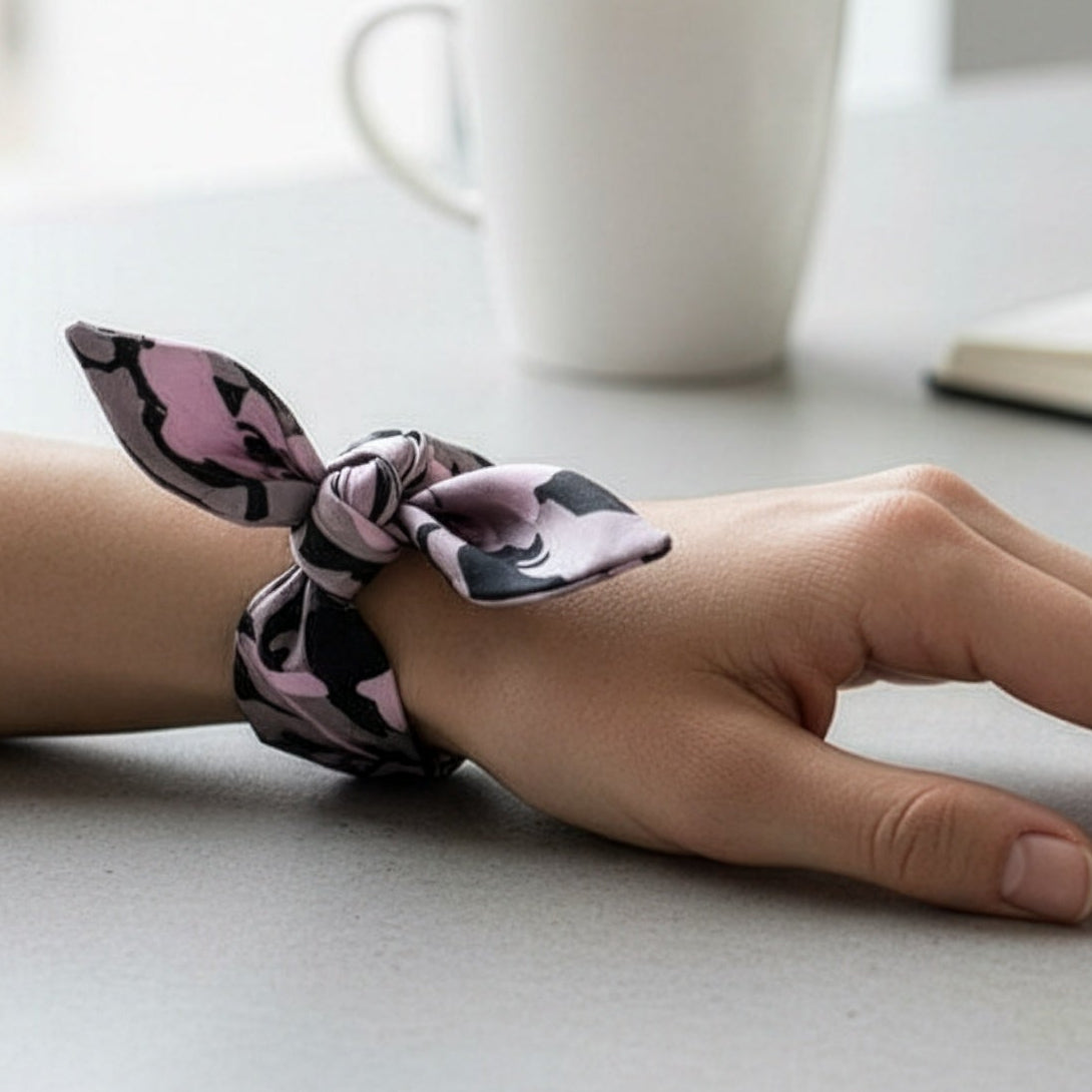 A close-up of a person's wrist wearing a satin bow headband with a pink, black, and white abstract pattern, set against a blurred background with a white mug.