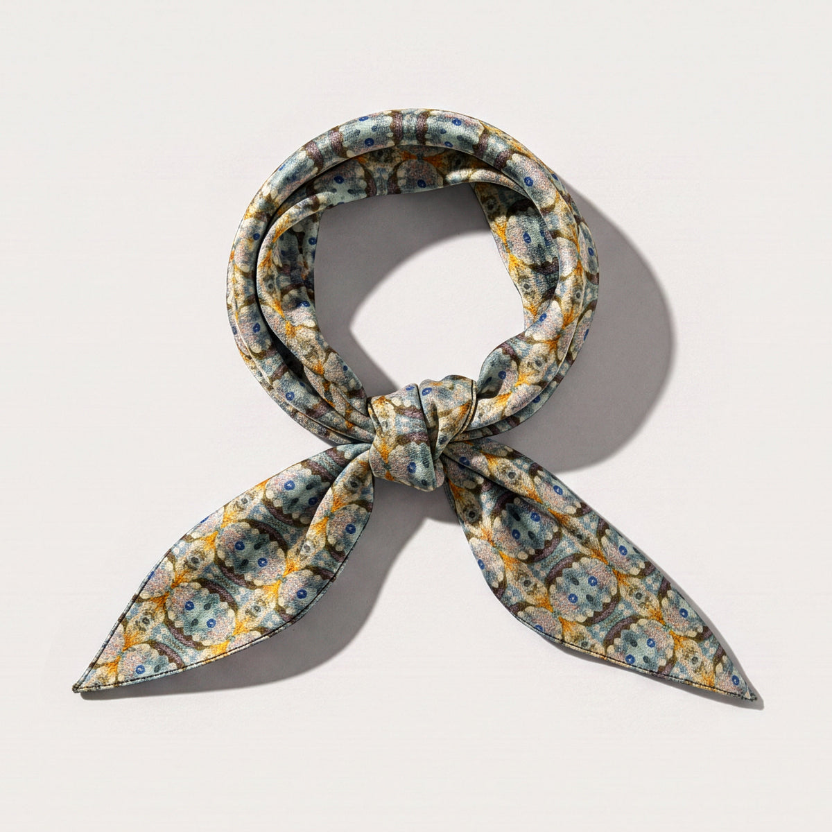 A patterned silk scarf tied in a loop with two pointed tails, featuring a yellow, blue, and gray floral design, set against a plain off-white background.