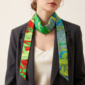 A woman wearing a black blazer and cream top models a vibrant silk scarf with a green, blue, and red abstract pattern against a plain beige background.