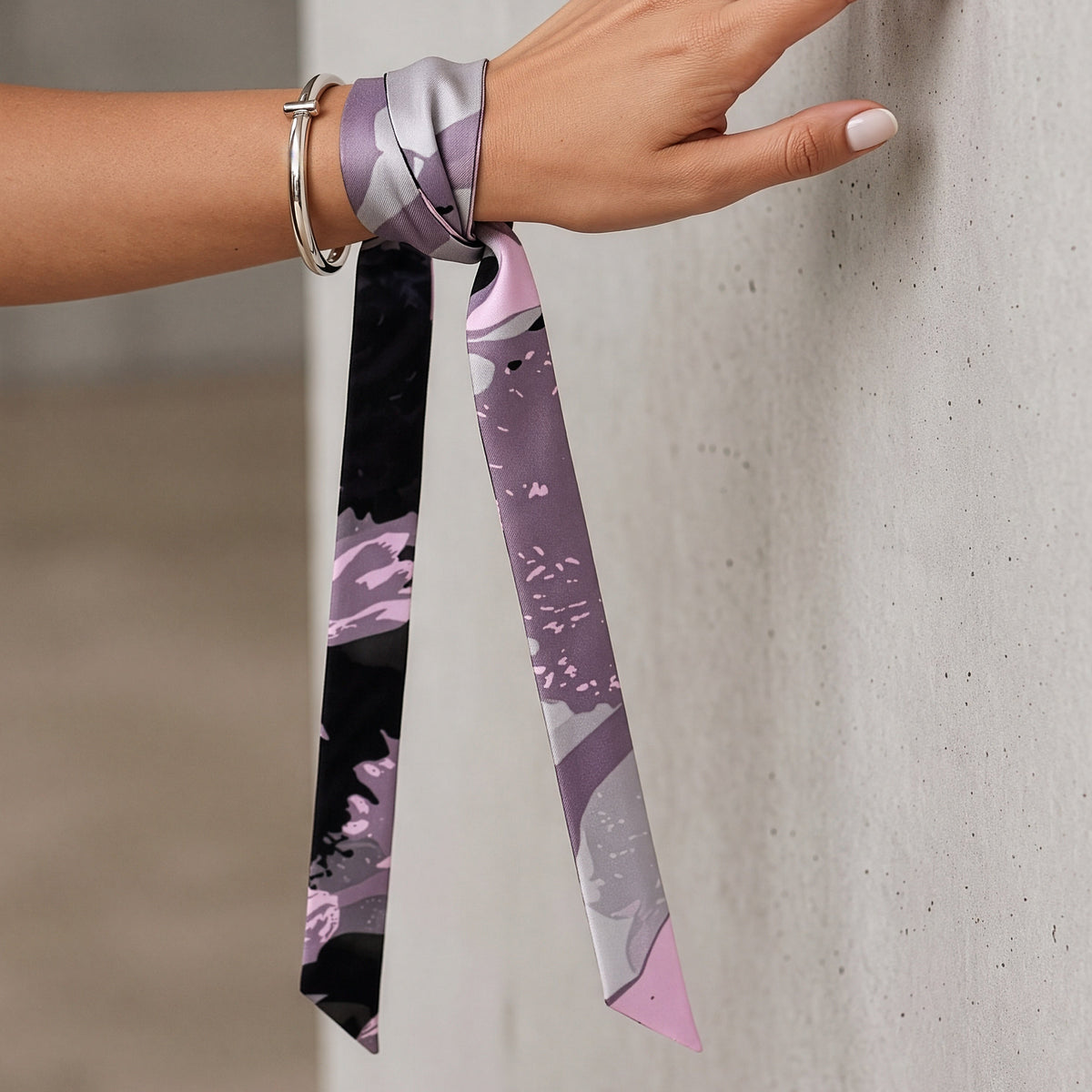 A close-up of a person's hand and wrist wearing a patterned silk scarf tied around the wrist, with the scarf hanging down against a textured concrete wall.