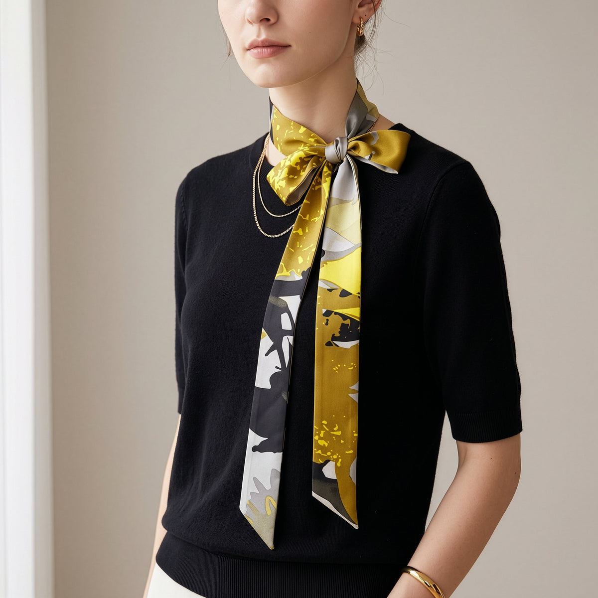 A woman wearing a black short-sleeve knit top and a patterned silk neck scarf in yellow, gray, and white, styled with gold necklaces and a gold bracelet.