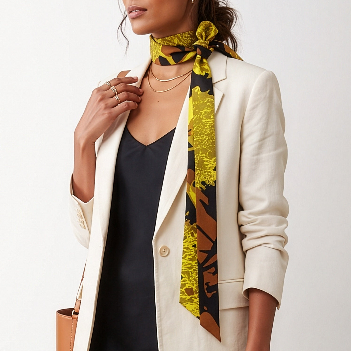 A woman models a stylish outfit consisting of a cream blazer, a black dress, a yellow and black patterned scarf, and black slide sandals, standing in front of a large white cylindrical structure.