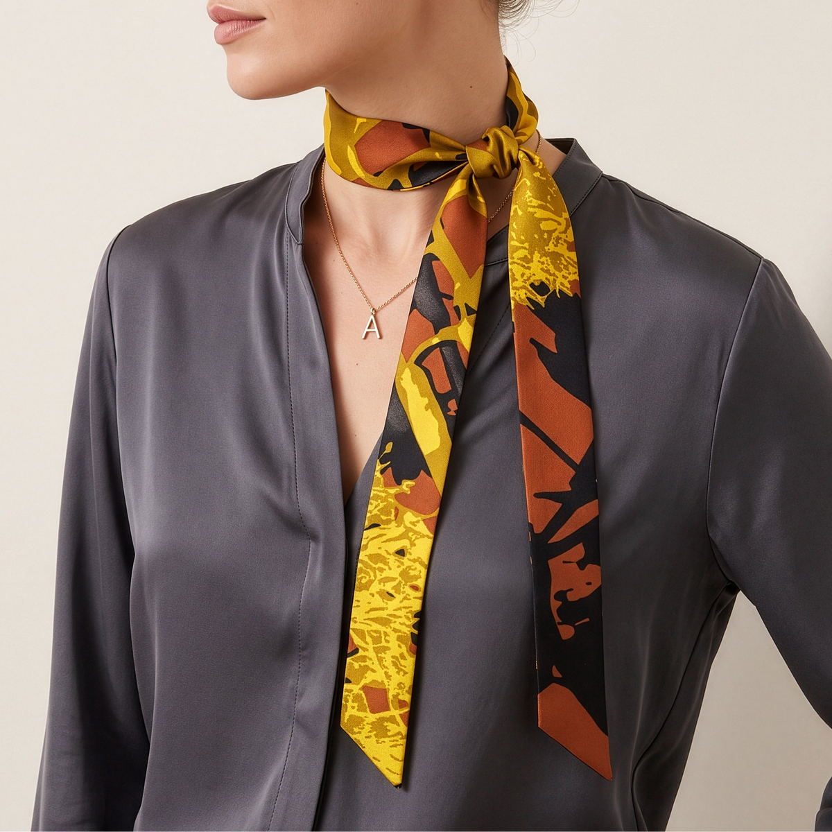 A woman wearing a dark gray satin blouse and a yellow, orange, and black patterned silk neck scarf, photographed against a warm off-white studio background.