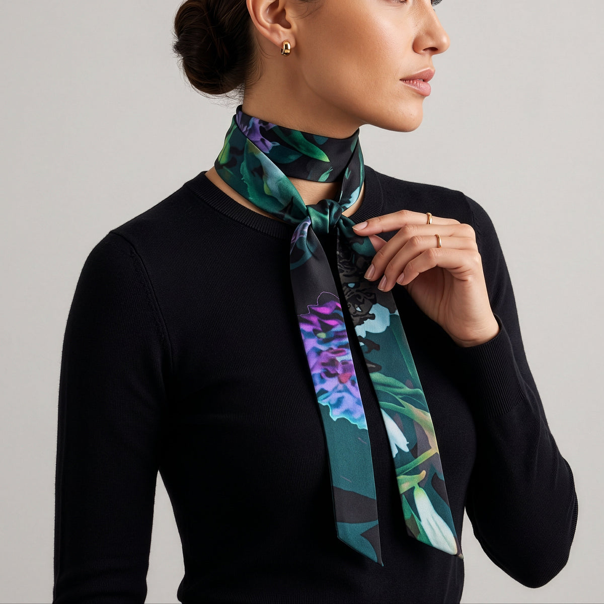A woman wearing a black long-sleeve top and a patterned silk neck scarf with green, purple, and blue floral details, posed against a neutral light gray background.