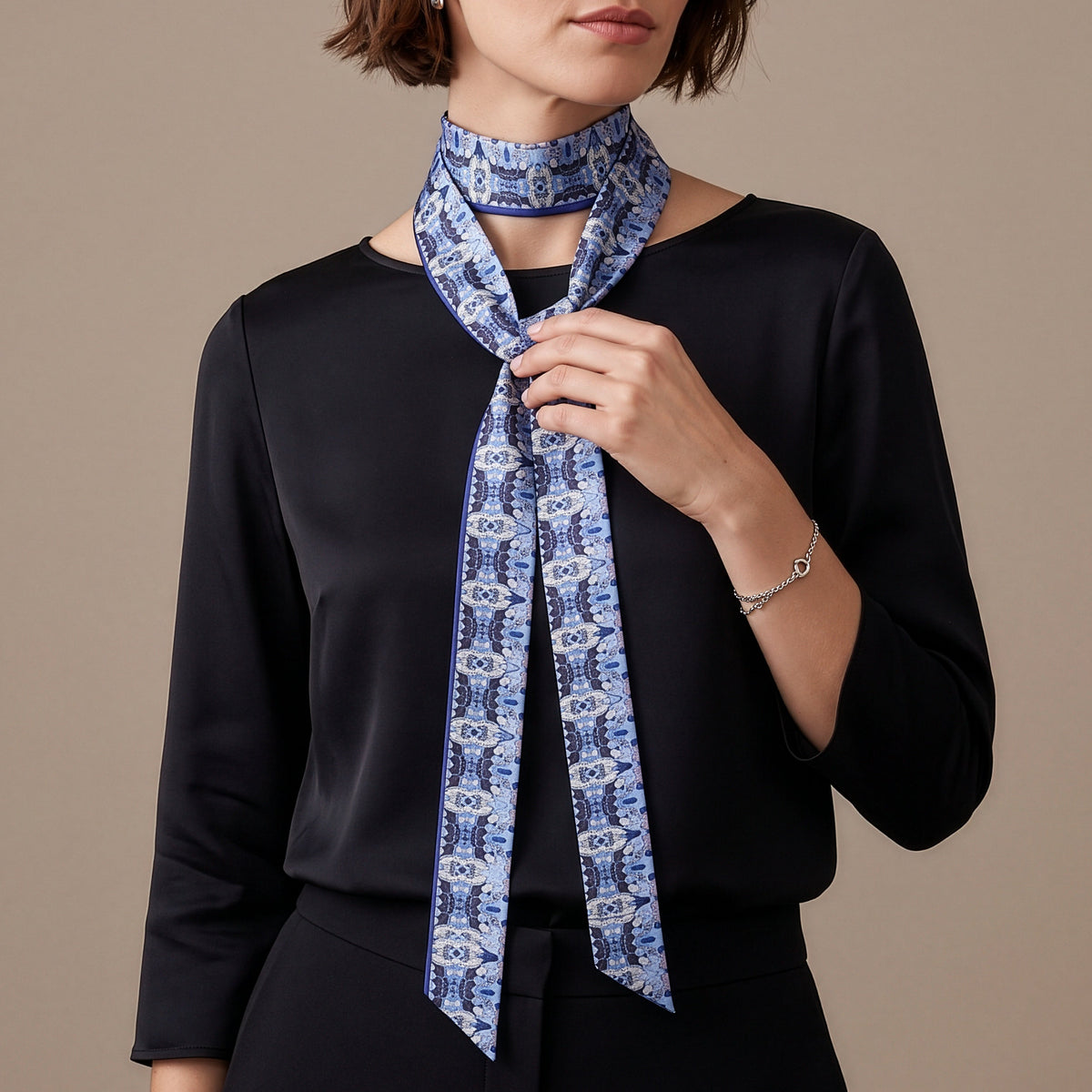 A woman with short brown hair models a blue and white patterned silk neck scarf tied around her neck, wearing a black long-sleeve top against a solid beige background.