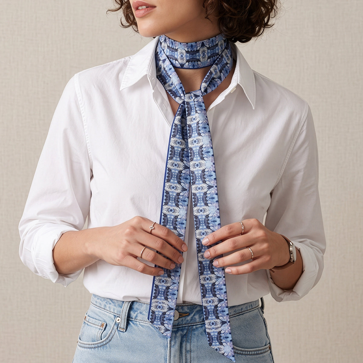 A woman wearing a white button-down shirt and light blue jeans, styled with a blue and white patterned silk neck scarf tied around her neck.
