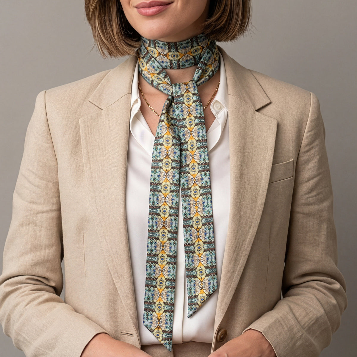 A woman with a short brown bob haircut models a patterned neck scarf tied around her collarbone, styled with a beige blazer and white shirt against a solid gray studio background.