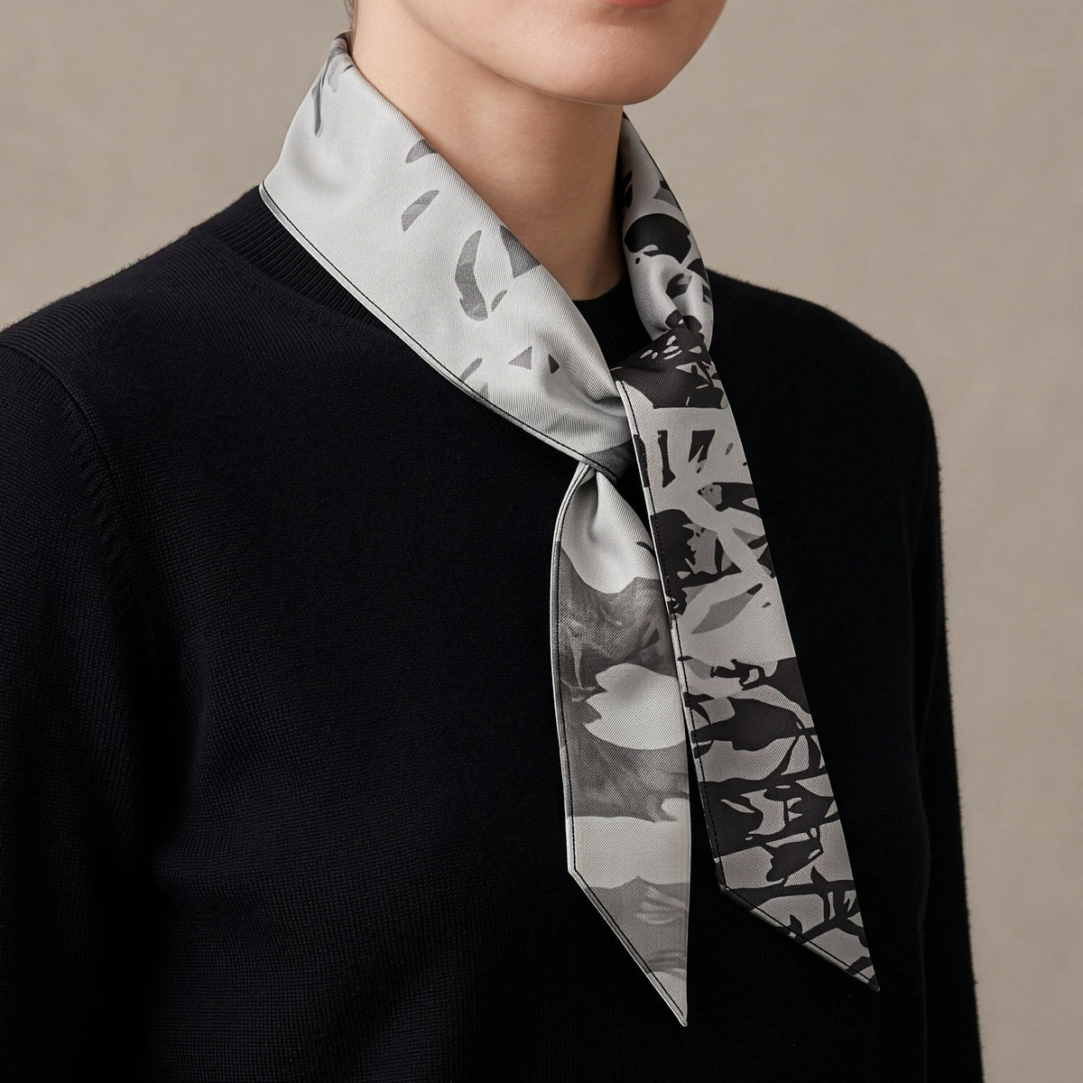 A close-up of a model wearing a black knit sweater and a gray and black abstract patterned silk neck scarf tied around the collar.