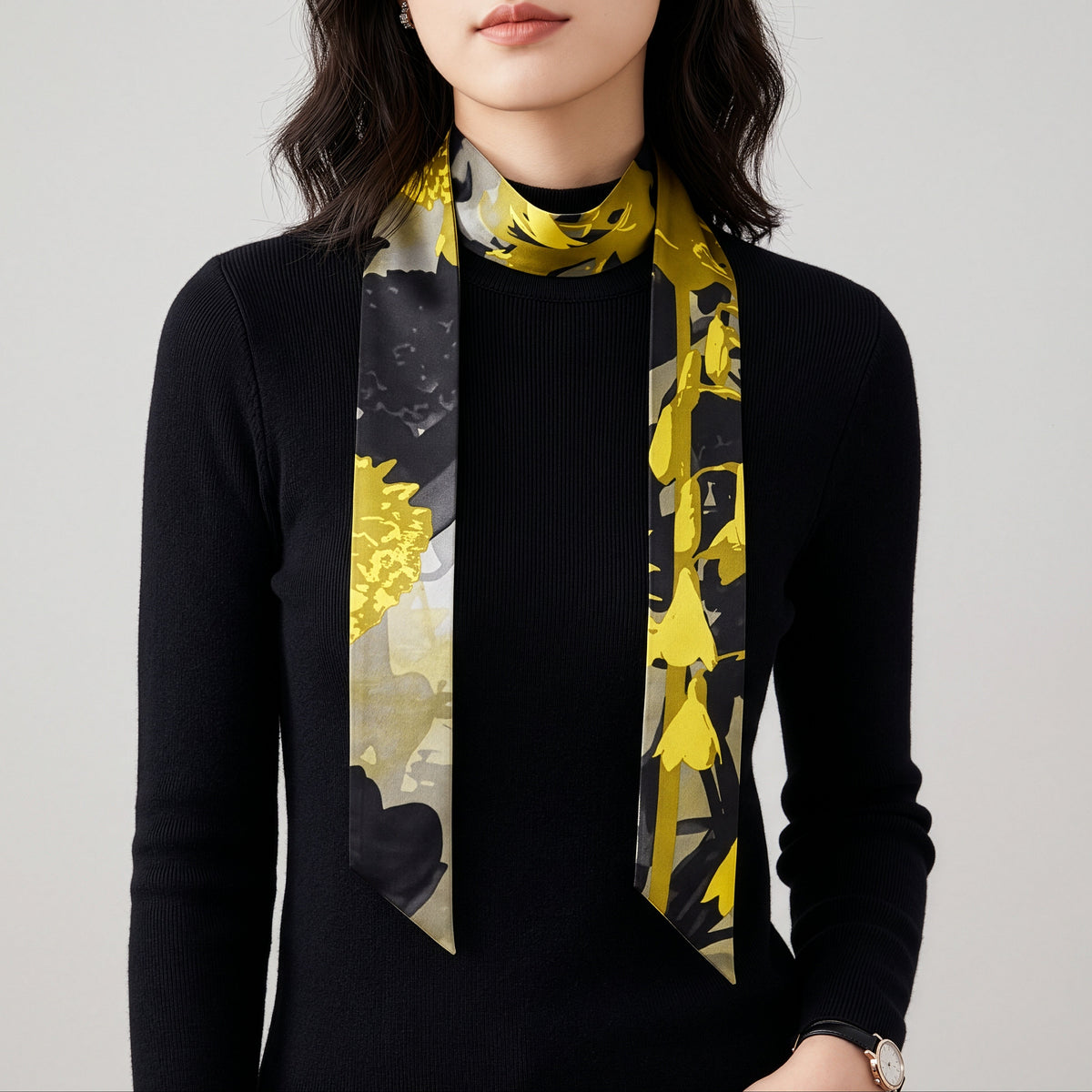 A woman wearing a black ribbed turtleneck sweater and a patterned silk scarf. The scarf features a bold yellow floral print over a black and gray background, styled around her neck against a plain light gray studio backdrop.