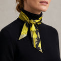 A woman wearing a black turtleneck sweater and a yellow and black patterned silk neck scarf, photographed against a neutral light gray background.