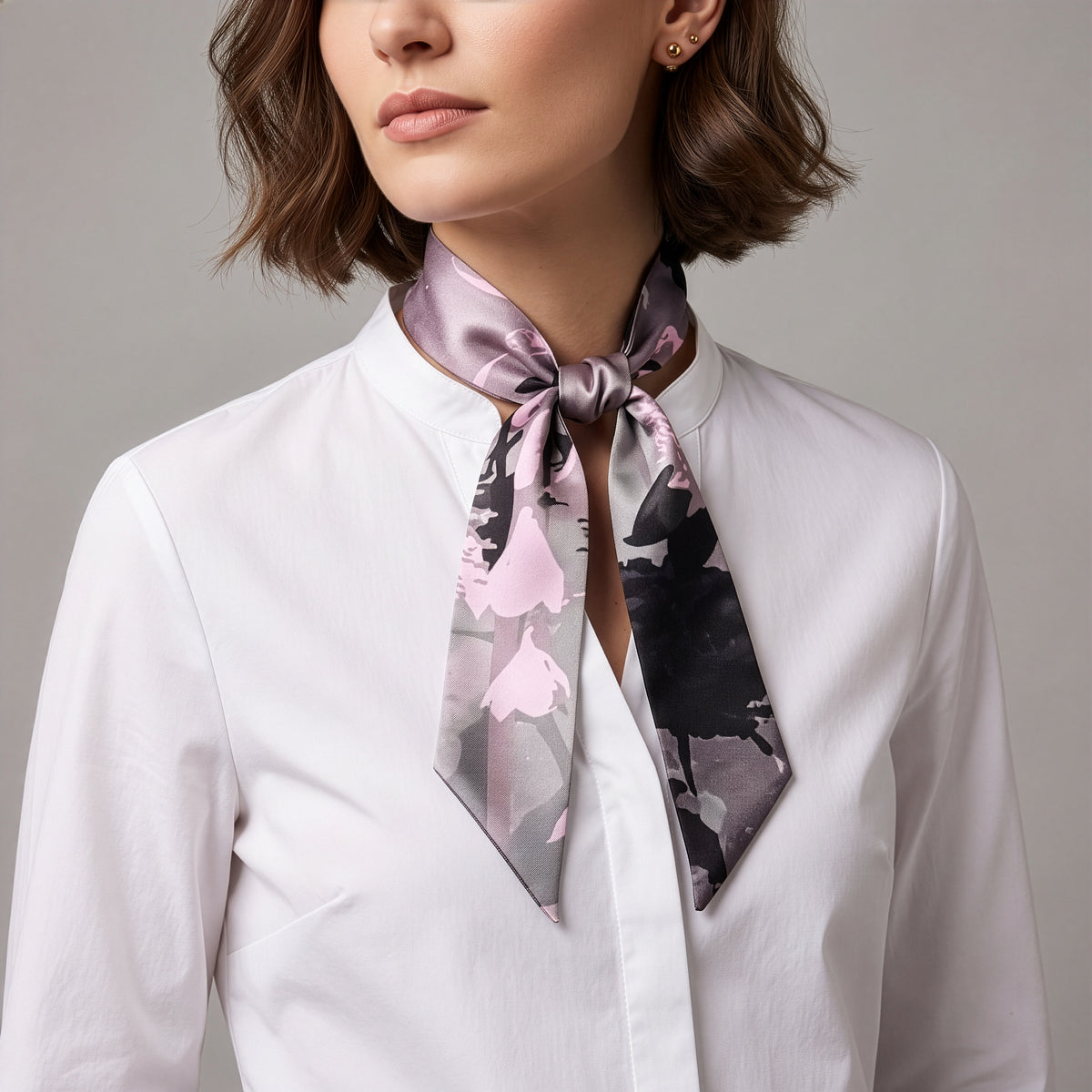 A close-up of a woman wearing a white collared blouse and a satin neck scarf with a pink, gray, and black abstract floral pattern, tied in a knot at the collar.