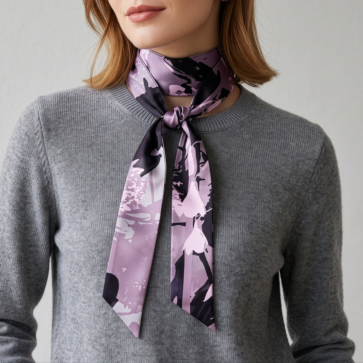 A woman wearing a gray knit sweater and a purple and black abstract-patterned silk scarf tied around her neck, photographed against a neutral light gray background.