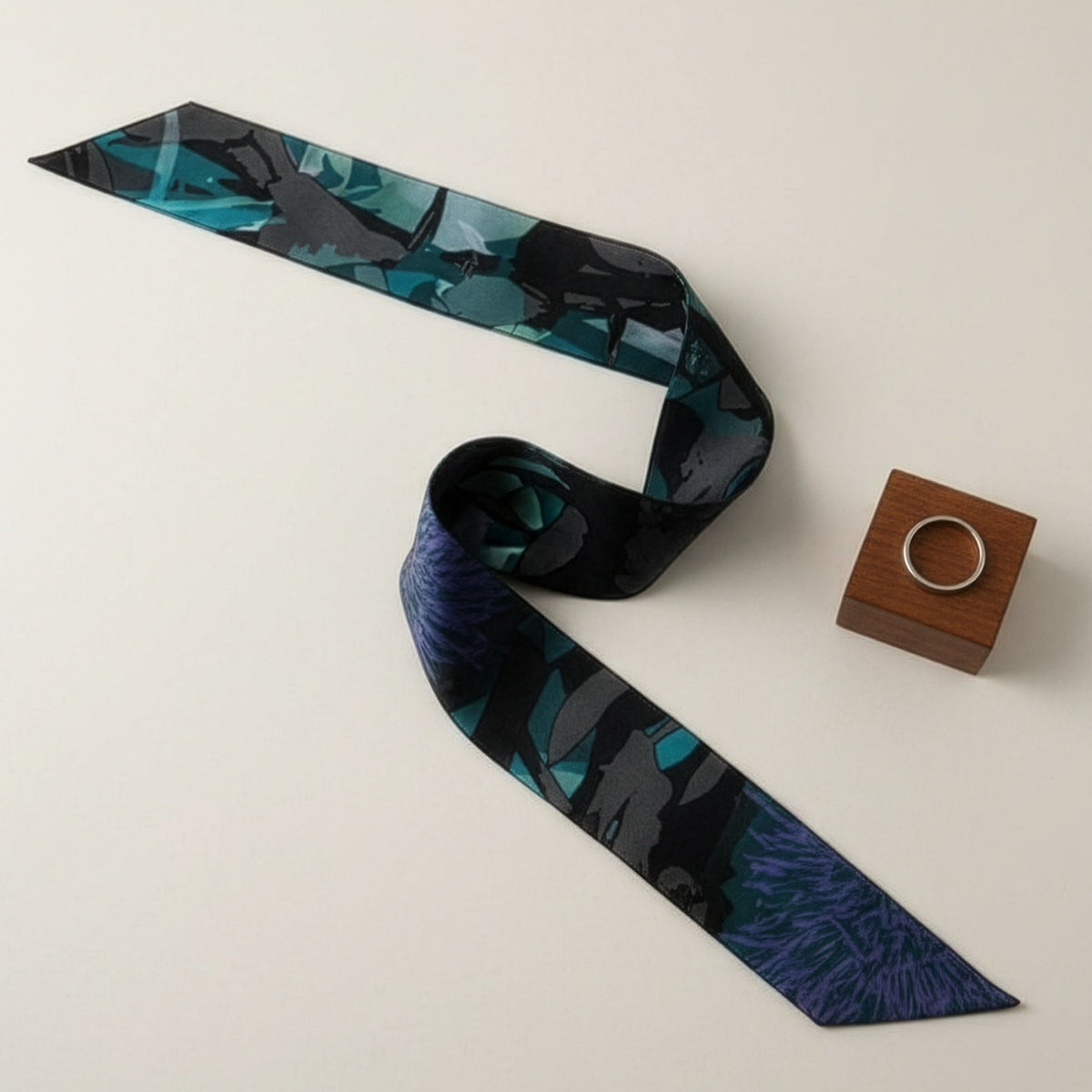 A slim necktie with a dark blue and black tropical leaf pattern, displayed on a light beige background next to a small wooden box holding a silver ring.